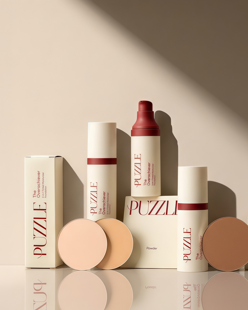 A set of makeup products from the brand PUZLE, including powder foundations, liquid foundation, and matching boxes, arranged on a reflective surface with a warm beige background and dramatic diagonal shadows.